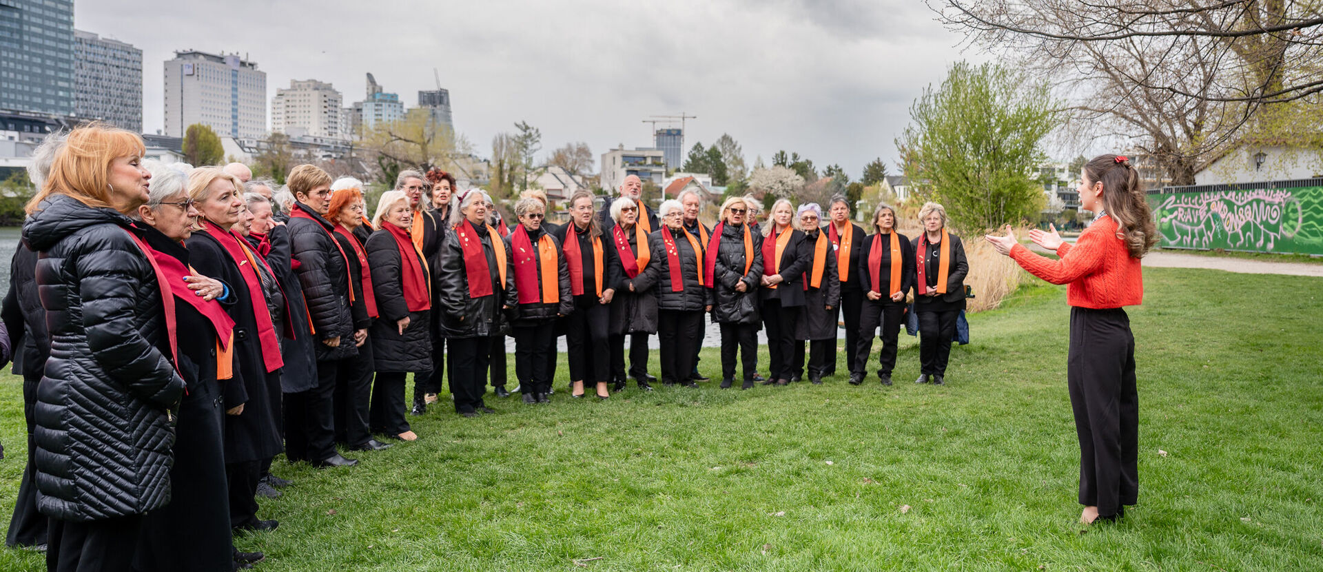 Der 1. Wiener Gemeindebauchor steht mit Chorleiterin Nelli Goncharuk auf einer Wiese und probt.
