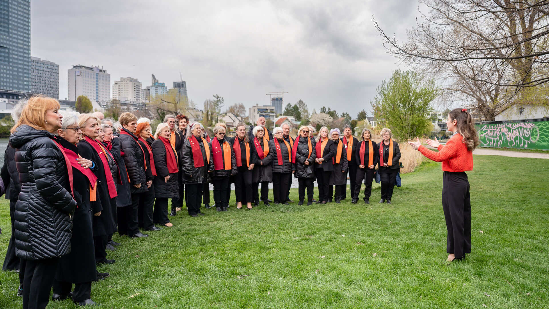 Der 1. Wiener Gemeindebauchor steht mit Chorleiterin Nelli Goncharuk auf einer Wiese und probt.