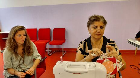 Zwei Frauen sitzen an einem Tisch mit einer weißen Singer-Nähmaschine, eine Frau hält ein Patchwork-Quiltstück in den Händen, im Hintergrund rote Stühle vor weißer Wand