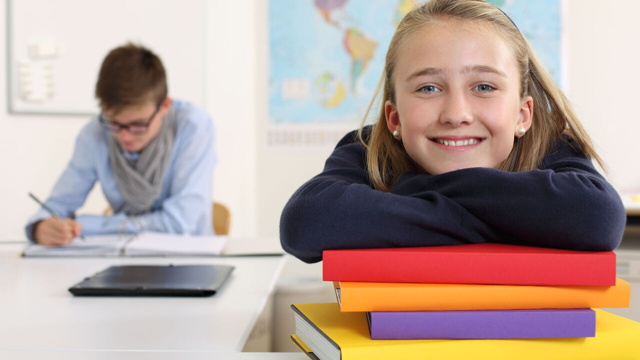 Zwei Schüler*innen. Die Person im hintergrund schreibt in eine Mappe, die Person im Vordergrund lehnt auf einem Stapel Bücher und lächelt in die Kamera