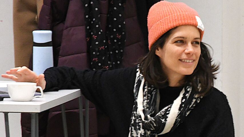 eine Person hockt auf dem Boden und ist an einem Tisch abgestützt, sie lächelt freundlich an der Kamera vorbei. Sie ist mit Jans, schwarzem Pulli und einer lachsfarbenen Wollhaube bekleidet und hat einen breiten schwarzweißen Schal umgehängt