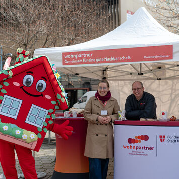 Ein Informationsstand von wohnpartner mit einer Mitarbeiterin, einem Mitarbeiter und dem Maskottchen WohnaLisa