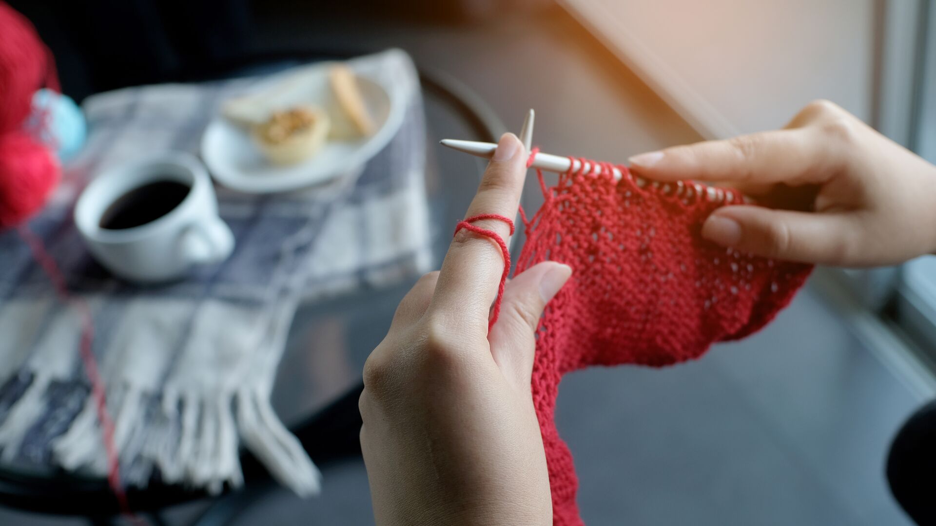 Zwei Frauenhände, die mit roter Wolle stricken. Im Hintergrund sieht man auf einer karierten Decke eine Tasse Kaffee und einen Teller mit Gebäck.