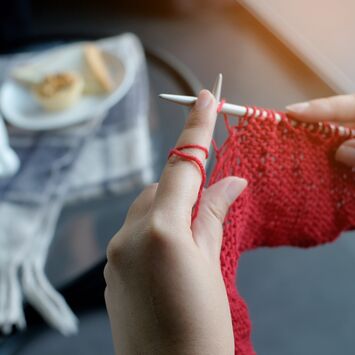 Zwei Frauenhände, die mit roter Wolle stricken. Im Hintergrund sieht man auf einer karierten Decke eine Tasse Kaffee und einen Teller mit Gebäck.