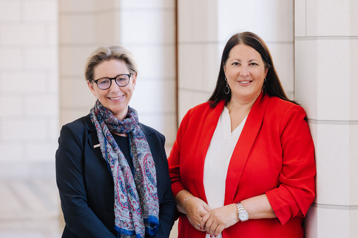 Zwei Frauen, Karin Ramser und Kathrin Gaál, stehen in einem Gang des Rathauses und lächeln in die Kamera. Beide sind businessmäßig mit Blazern gekleidet.