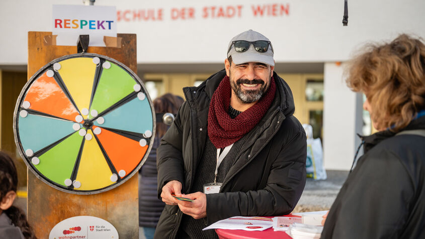 Ein freundlich lächelnder Mitarbeiter von wohnpartner, erkennbar an seinem Patch, steht neben einem Glücksrad . Er ist offensichtlich für kühleres Wetter gekleidet.
