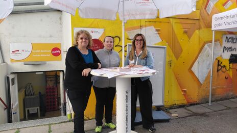 Drei Frauen stehen an einem Stehtisch mit Sonnenschirm vor einer gelb bemalten Wand mit Plakaten