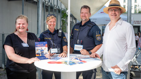 Zwei Polizisten in Uniform und zwei Zivilpersonen stehen an einem Stehtisch mit Flyern und lächeln in die Kamera.