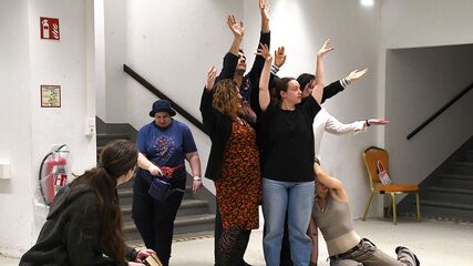 Eine Person sitzt von der Kamera abgewandt auf einer Bank, daneben stehen einige Personen und strecken ihre Arme in die Luft. Eine Person sitzt seitlich angelehnt an die Gruppe davor. Eine weitere Person geht hinter der Gruppe vorbei. e 