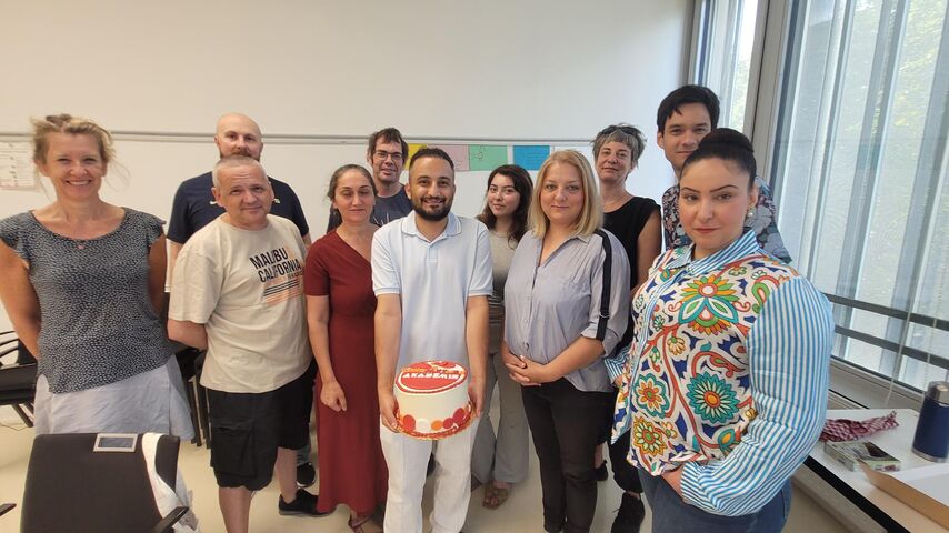 Gruppe von elf Personen in einem hellen Raum, eine Person hält einen runden Kuchen mit roter Dekoration