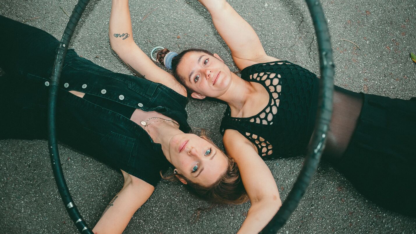 Zwei junge Frauen liegen Kopf neben Kopf in gegengesetzter Richtung auf dem Asphalt und blicken in die Kamera. Sie sind mit schwarzen Shirts und schwarzen Leggings bekleidet und halten einen Hulahoop-Ring in ihren Händen.